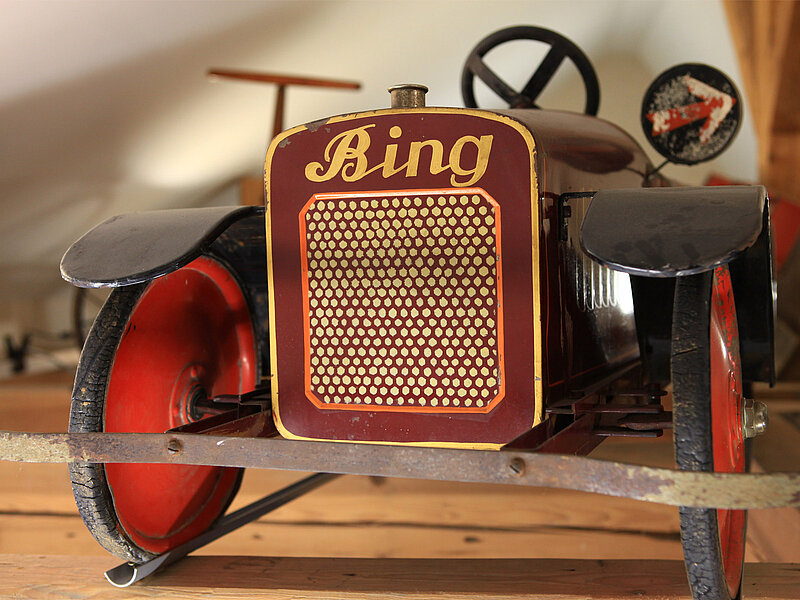 Ein altes, rotes Spielzeugauto mit der Aufschrift "Bing" auf der Vorderseite. Das Auto hat runde Räder und eine einfache Steuerung. Es ist auf einem Holzregal platziert, umgeben von einer hellen, neutralen Umgebung.